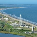aerial view of the coastal launch range of Wallops Flight Facility, showing a blue Atlantic Ocean on the right; white buildings along a tan coastline back up to a green, marshy landscape